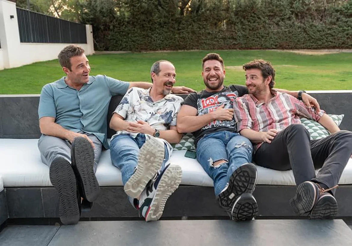 Four men sitting on an outdoor sofa, laughing with their feet toward the camera in a sunny yard setting