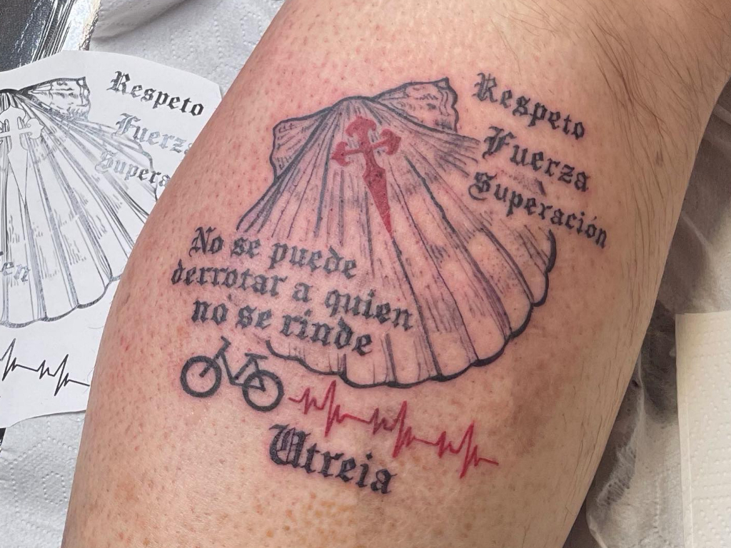 Shell tattoo featuring a red cross, Spanish mottoes about respect, strength, and perseverance, a bicycle, and a red heartbeat line with the name 'Atria'