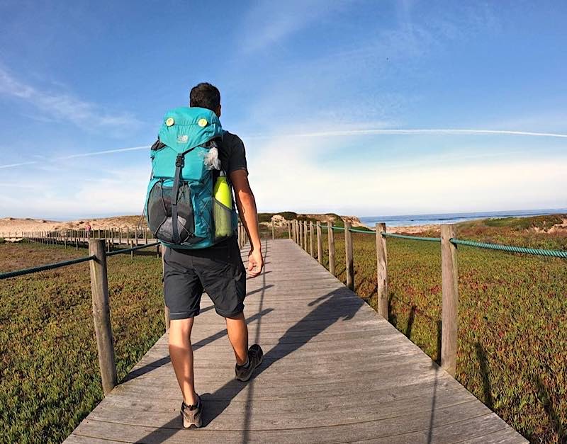 Person wearing a teal backpack walking along a wooden boardwalk toward the ocean on a sunny day with blue sky.
