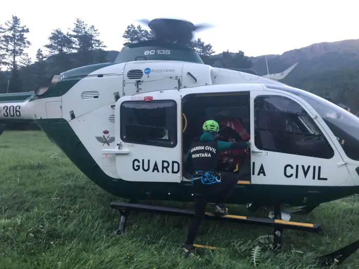Green-and-white Guardia Civil helicopter on a grassy field with the side door open; a rescuer in a bright helmet and safety harness climbs into the cabin.