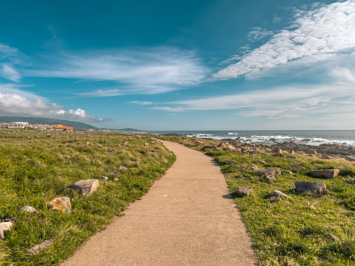 Paved coastal path winds through grassy, rocky shoreline toward the ocean under a blue sky with clouds.