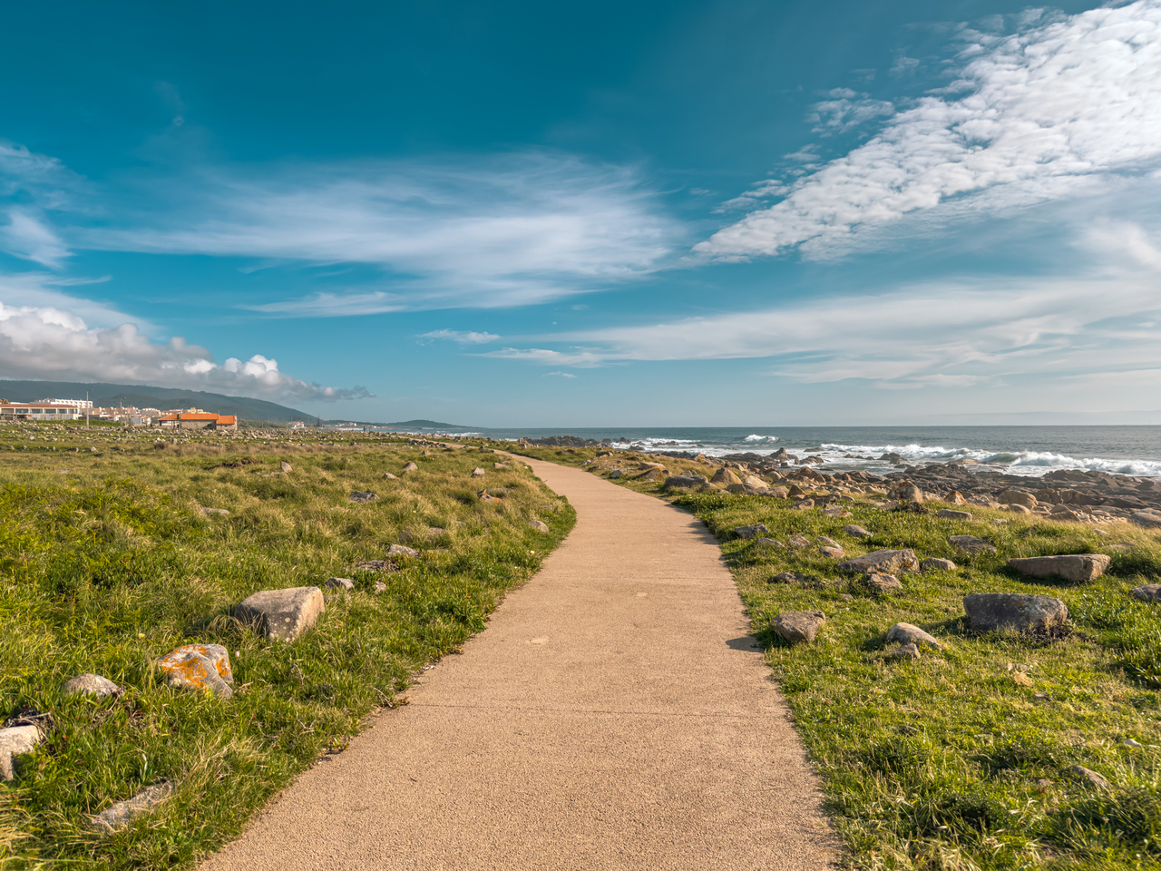 Paved coastal path winds through grassy, rocky shoreline toward the ocean under a blue sky with clouds.