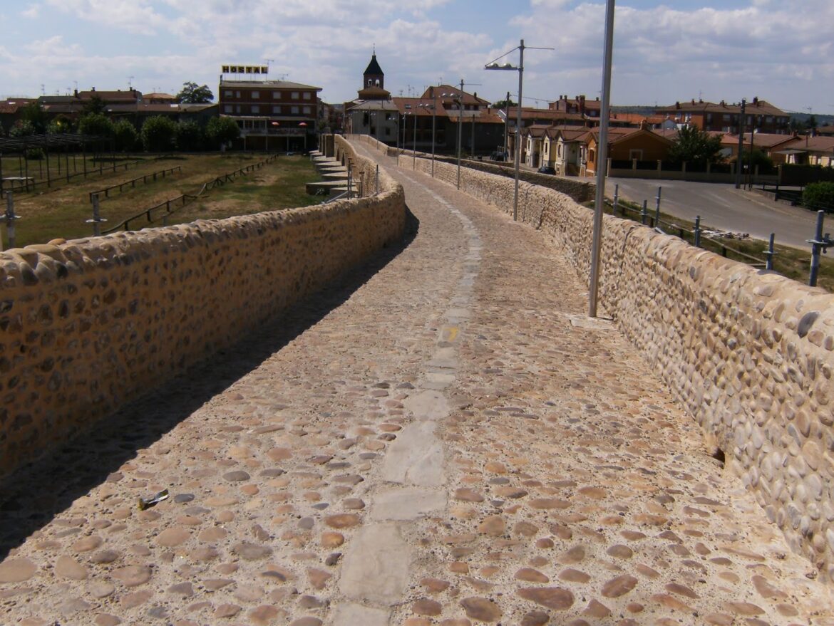 Curving cobblestone walkway on a historic stone bridge with low walls leading to a town square in the distance.