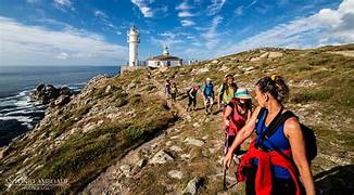 Hikers walk along a rocky coastal path toward a white lighthouse on a cliff, with the sea on the left and a blue sky above.