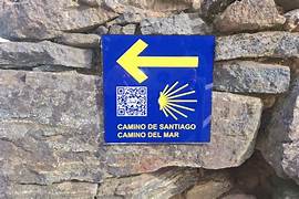 Blue sign with a yellow left arrow, a QR code, and a scallop shell indicating the Camino de Santiago (Camino del Mar) on a stone wall