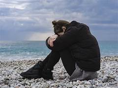 Person sits alone on a pebble beach, arms wrapped around their knees, looking down toward the sea under a cloudy sky.