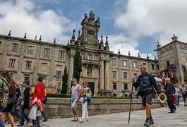 Tourists walking across a sunny plaza in front of a large historic palace with statues along the roofline.