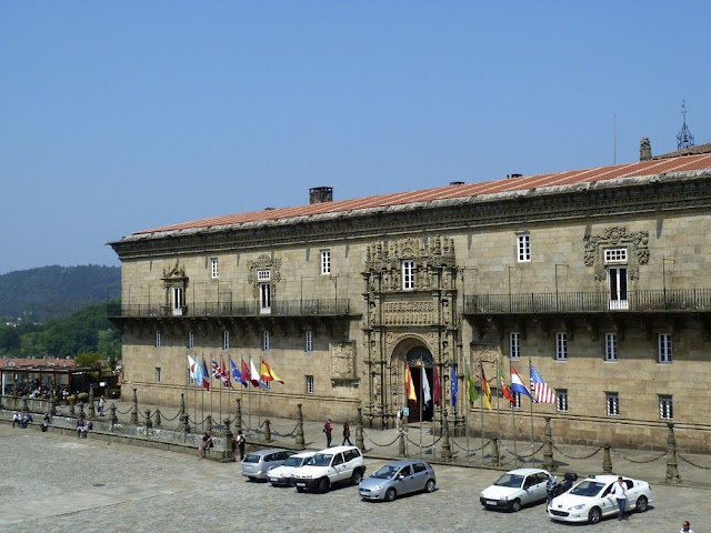 Historic stone building with ornate doorway, row of flags along the front, and parked cars in a cobblestone plaza.