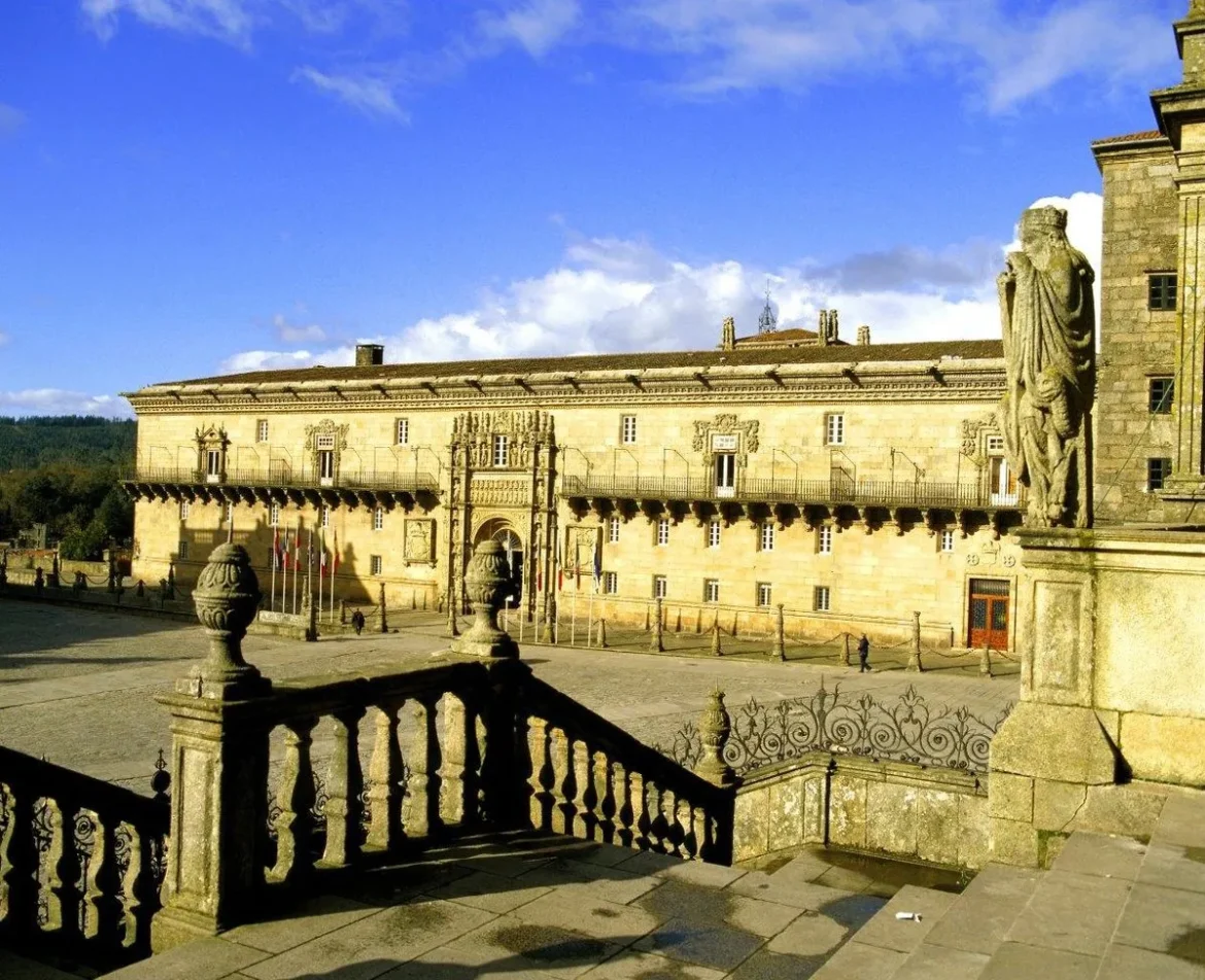 Sunlit historic stone building with ornate doorway in a wide plaza, seen from a terrace with decorative railing in the foreground.