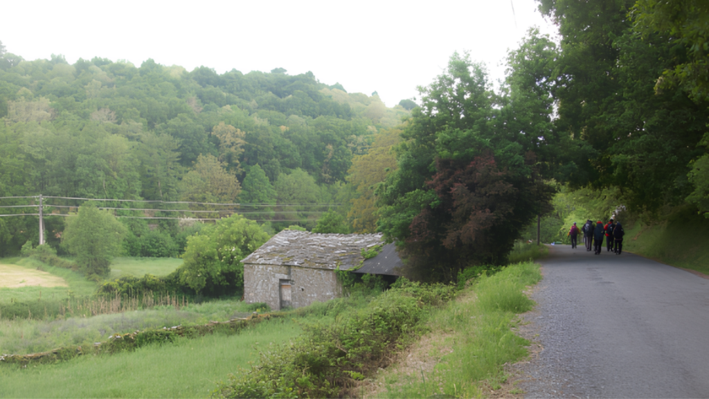 Group of hikers walking along a rural road beside a small stone cottage in a green valley.