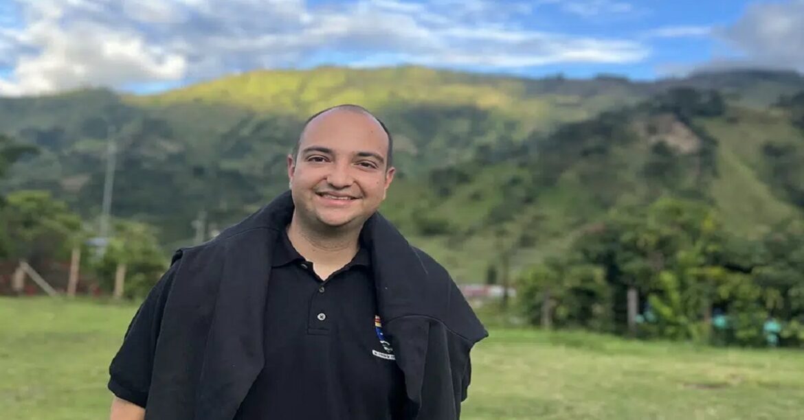 Smiling man in a black polo shirt with a dark jacket draped over his shoulders, standing in a grassy field with green hills and blue sky behind him.