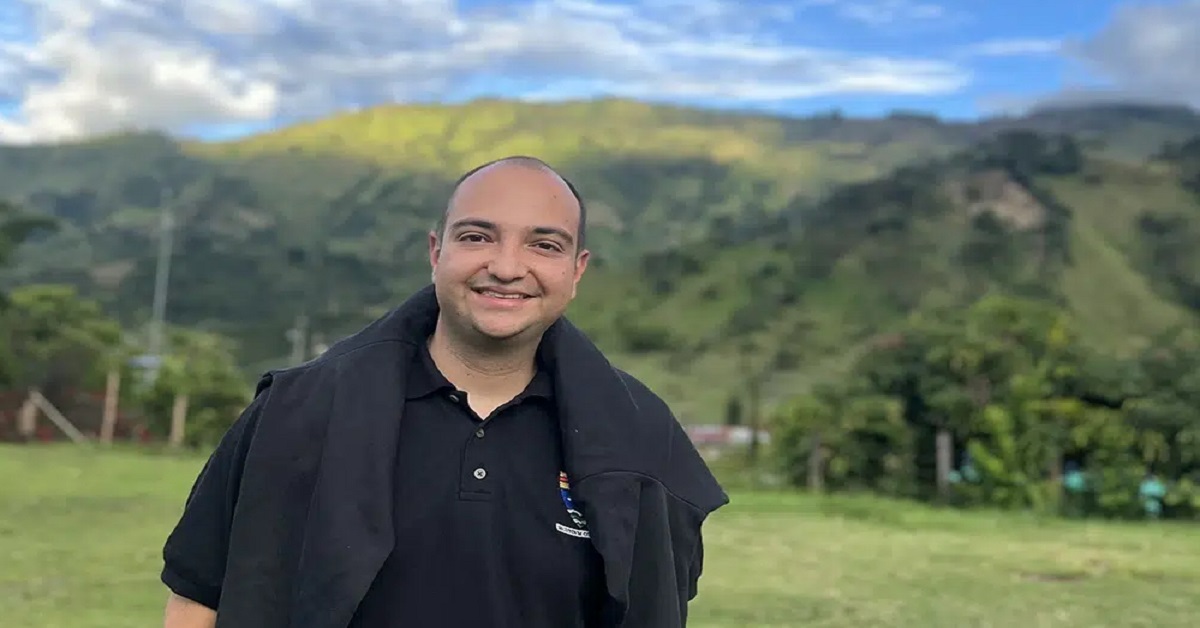 Smiling man in a black polo shirt with a dark jacket draped over his shoulders, standing in a grassy field with green hills and blue sky behind him.