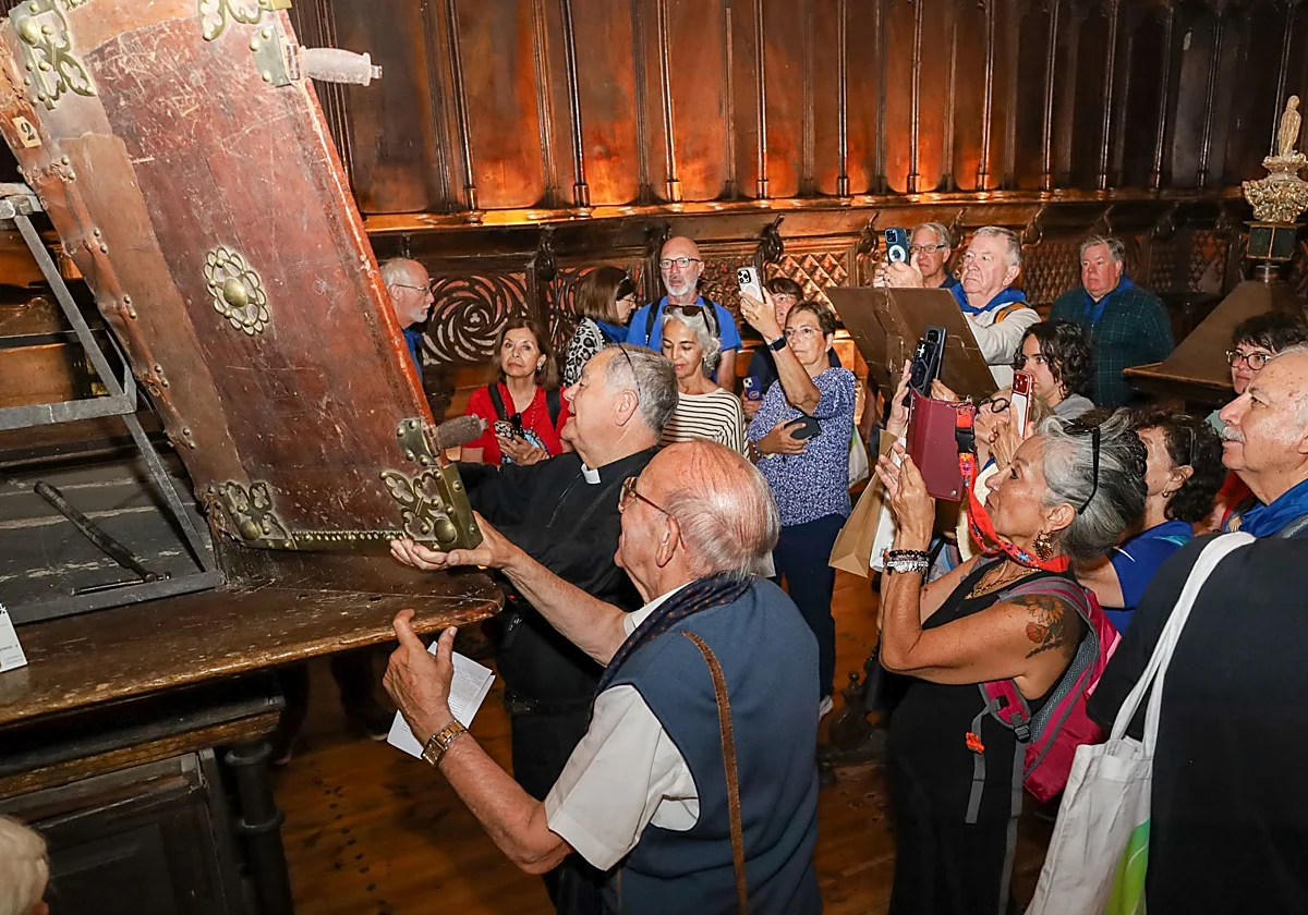 Priest and a group of tourists gather around a large ornate wooden chest as it is opened, with many people taking photos.
