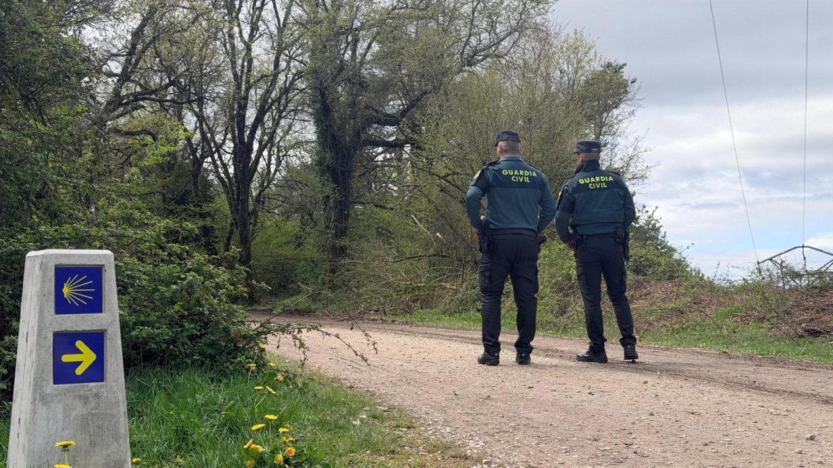 Two Guardia Civil officers stand on a dirt path in a wooded area, facing away from the camera, near a concrete milestone with blue signs and a yellow shell symbol and arrow to the right.