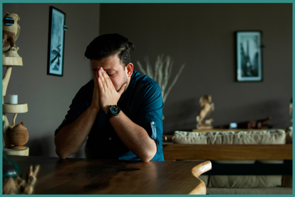 Man in a blue shirt sits at a wooden table with his face buried in his hands, looking distressed in a dim living room.