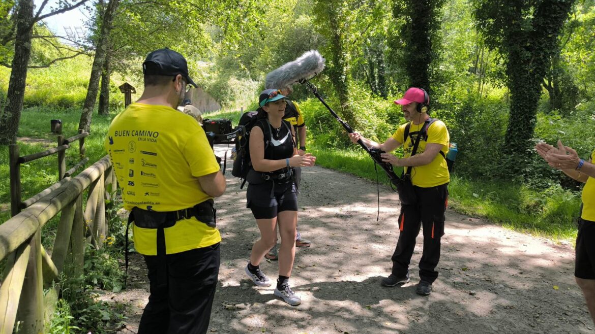A woman hiker with a backpack and harness is interviewed by a crew on a forest trail, while a man with a microphone boom stands nearby wearing a yellow shirt and cap.