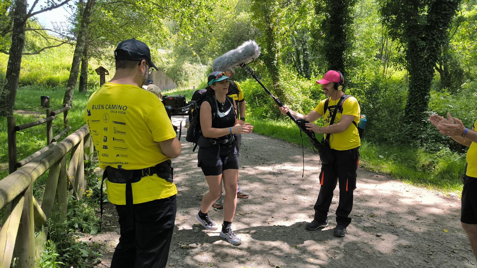 A woman hiker with a backpack and harness is interviewed by a crew on a forest trail, while a man with a microphone boom stands nearby wearing a yellow shirt and cap.