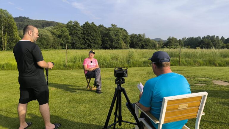 A man in a pink shirt sits on a chair outdoors while two crew members film him with a camera on a tripod in a sunny grassy field.