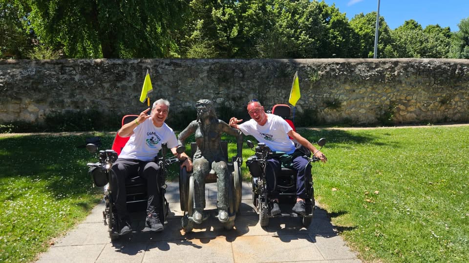 Two men in power wheelchairs pose on either side of a bronze statue in a sunny park, smiling at the camera with a stone wall and green lawn behind them.