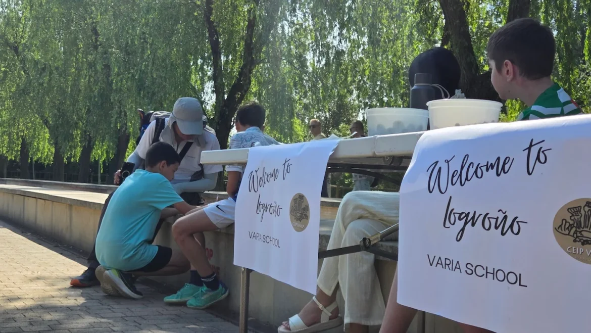 Outdoor scene of a welcome event at a school: people gather around a table with banners reading 'Welcome to' and 'VARIA SCHOOL', a man helps a young boy with something by the barrier.