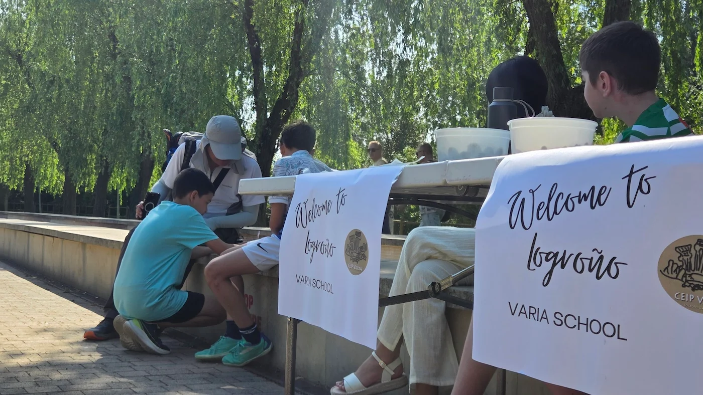 Outdoor scene of a welcome event at a school: people gather around a table with banners reading 'Welcome to' and 'VARIA SCHOOL', a man helps a young boy with something by the barrier.