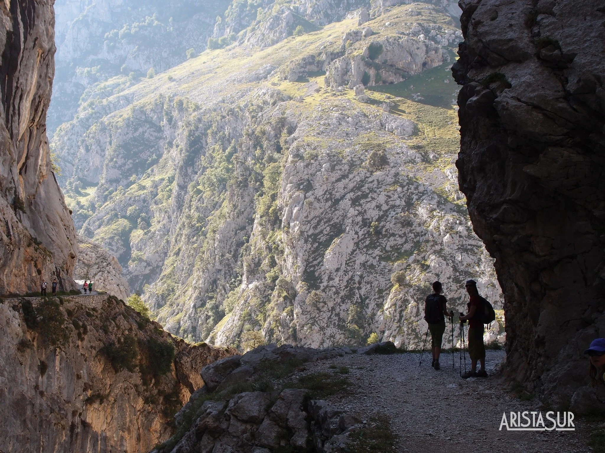 Wanderer mit Trekkingstöcken wandern entlang eines felsigen Canyon-Pfades zwischen hoch aufragenden Felswänden. Leichtes Sonnenlicht beleuchtet das Tal vor uns.