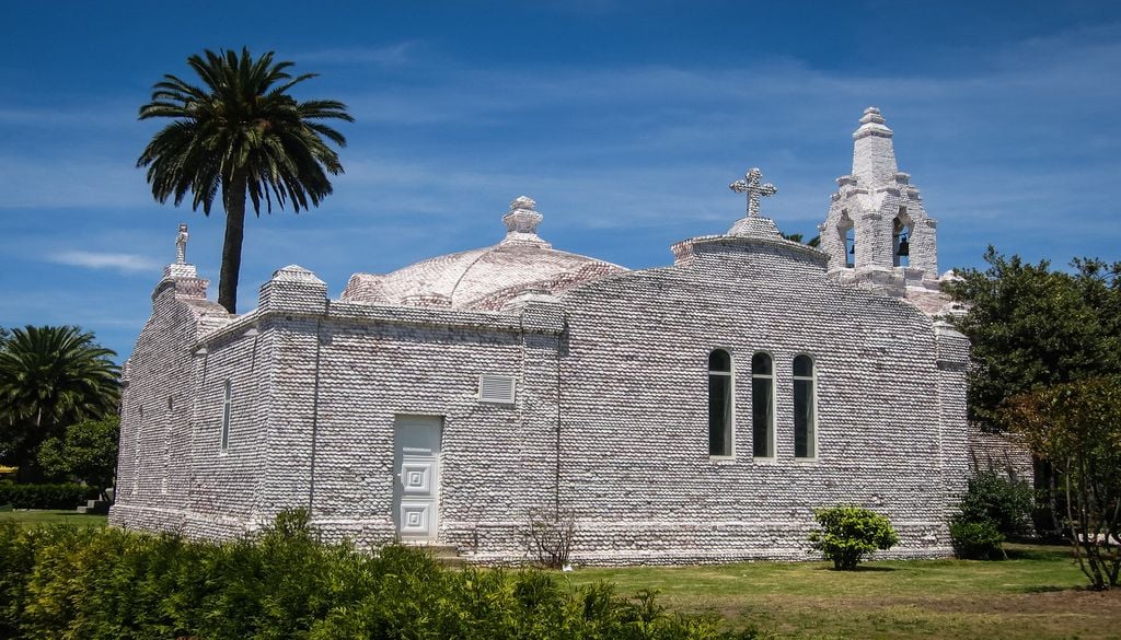 White stone chapel with arched windows and crosses on the roof, set in a sunny garden with palm trees nearby