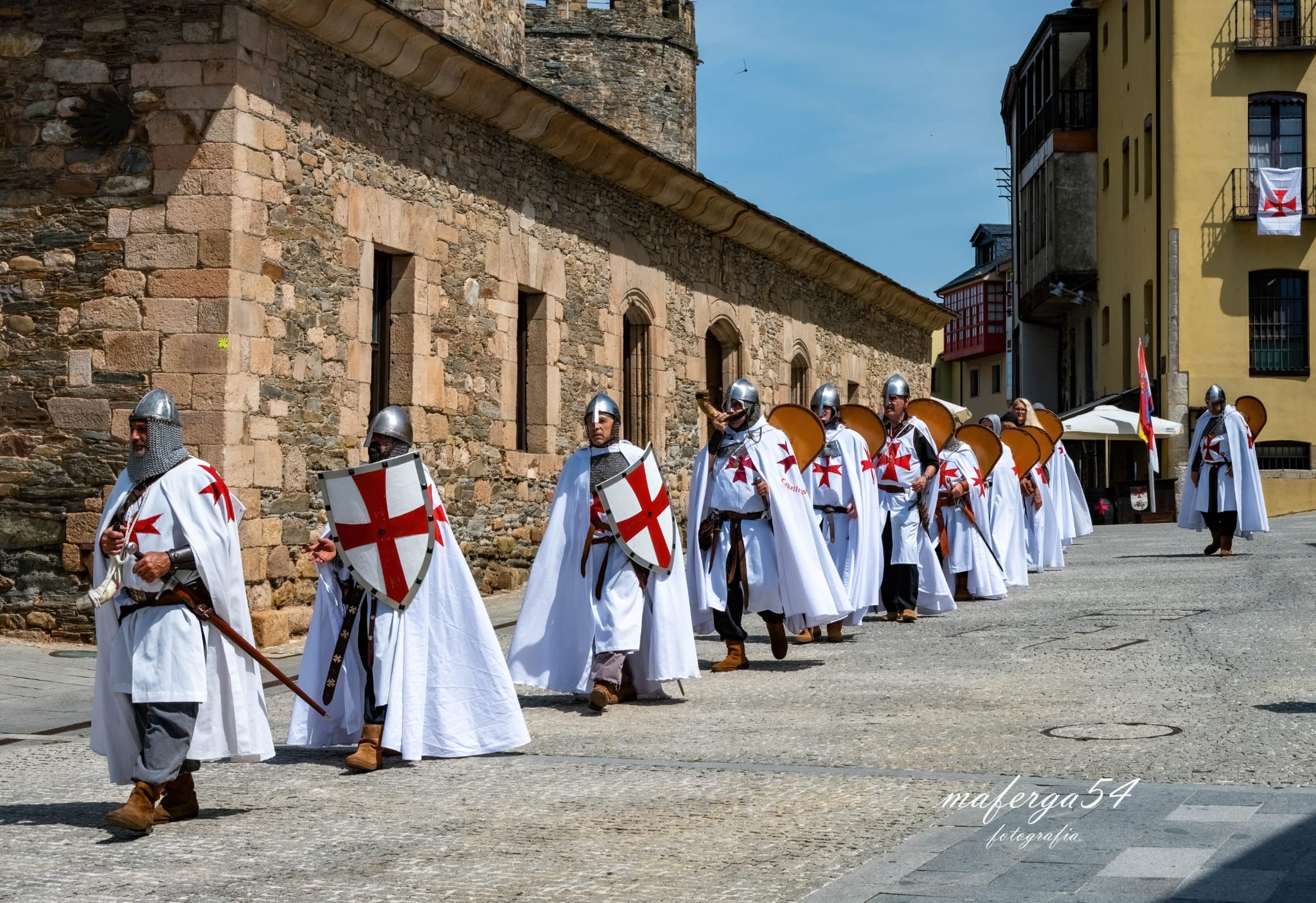 Line of medieval knights in white tunics with red crosses marching along a cobblestone street beside a historic stone wall.