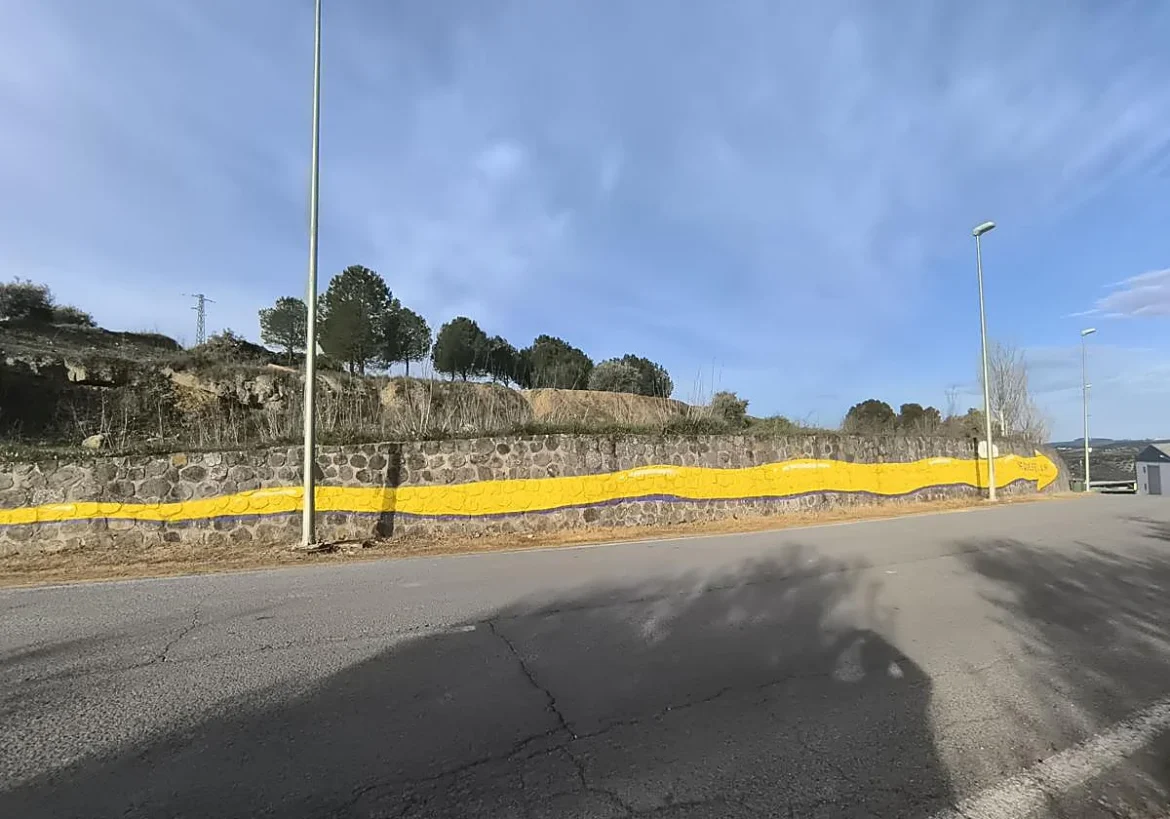 Stone wall along a road with a long yellow wavy mural under a blue sky, trees on a hill above the wall.