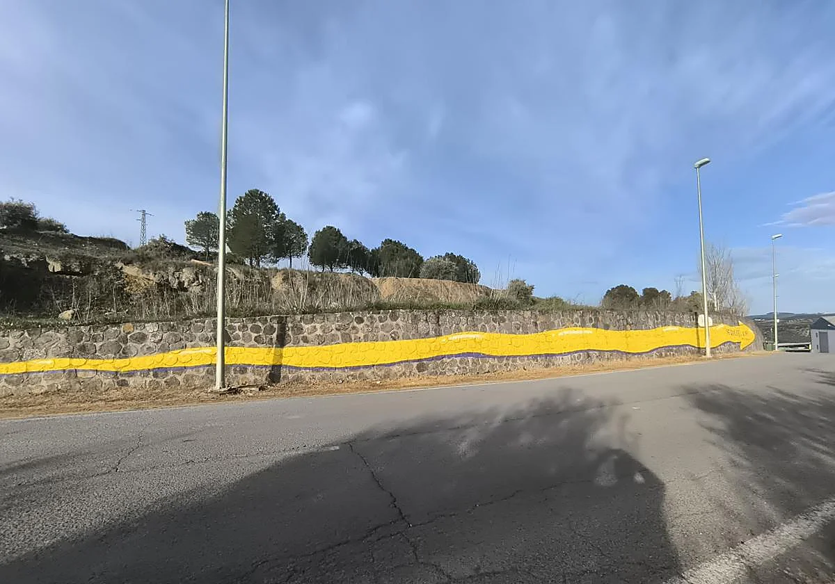 Stone wall along a road with a long yellow wavy mural under a blue sky, trees on a hill above the wall.