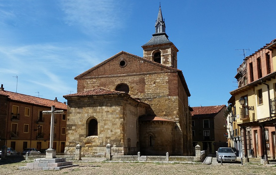 Stone church with a small bell tower in a town square, surrounded by brick buildings and a cobblestone plaza.