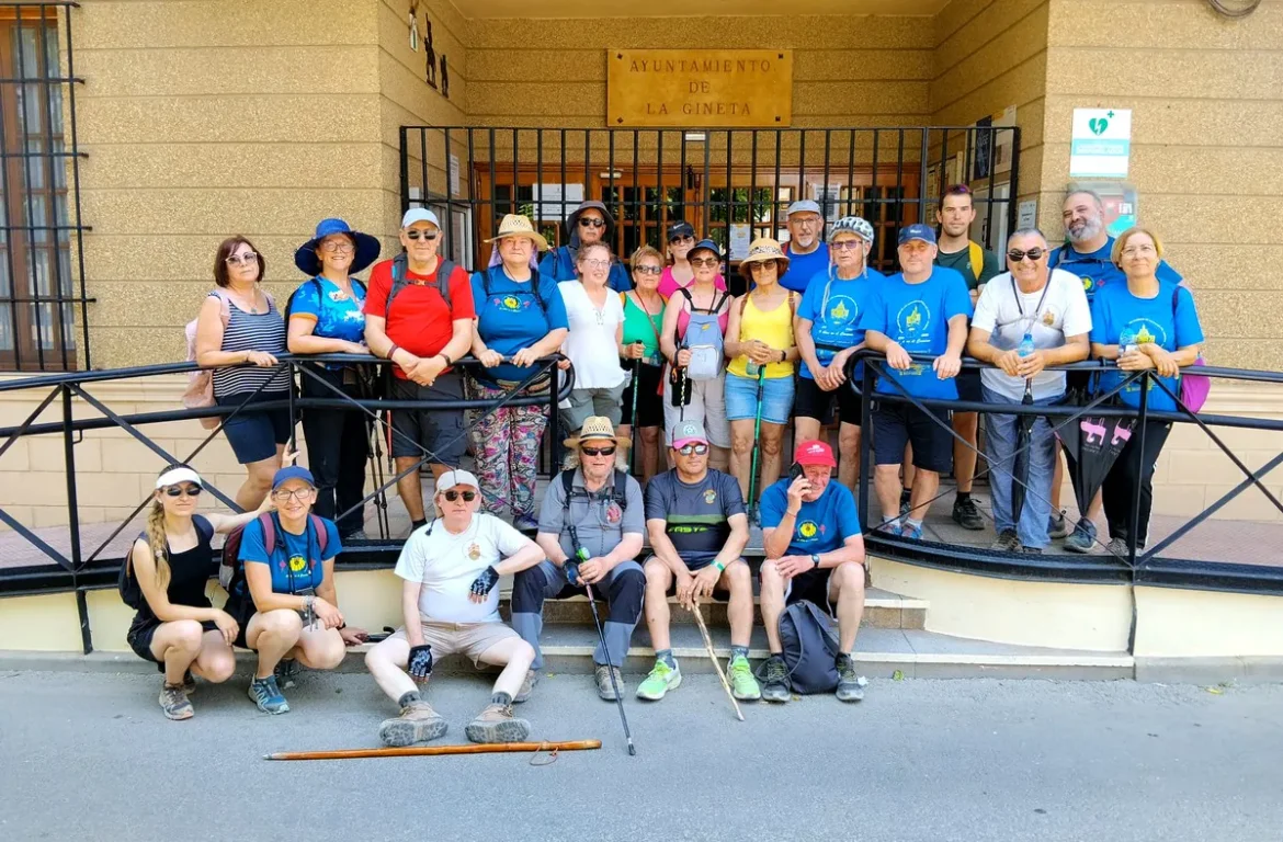 Group of hikers posing on the steps in front of the Ayuntamiento de La Gineta, wearing blue shirts and sun hats.