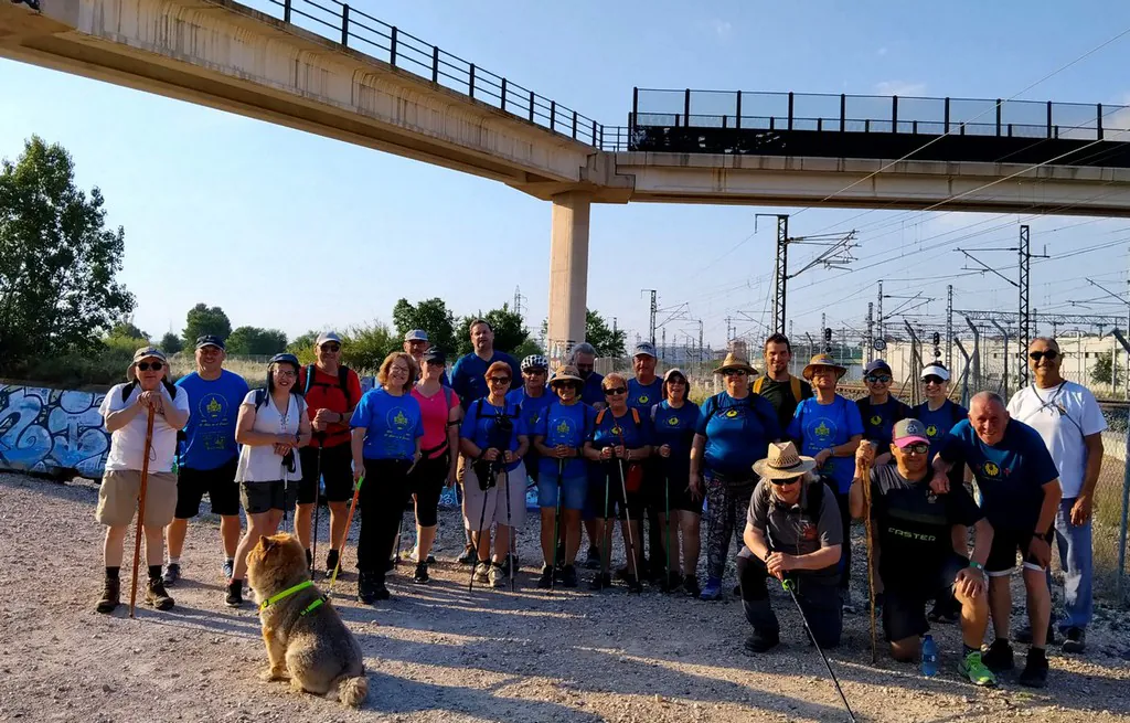 Group of hikers posing for a photo under a concrete railway overpass, with a dog sitting at the front wearing a bright collar.