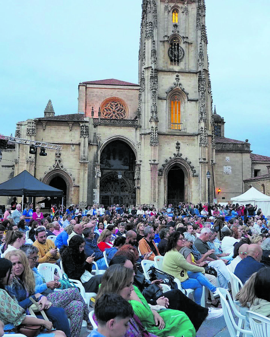 Crowd of people seated on white plastic chairs at an outdoor event in front of a Gothic church with a tall ornate tower and arched entrances.