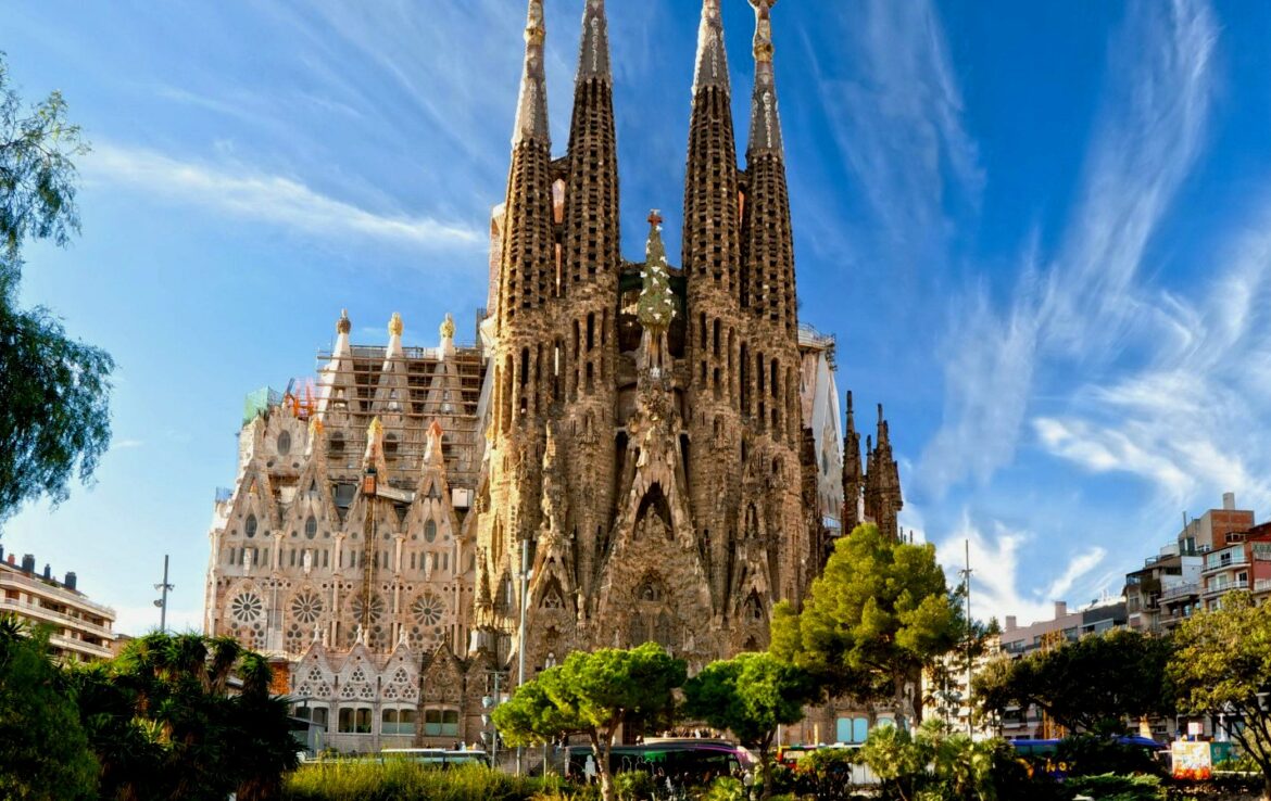 Sagrada Família basilica in Barcelona under construction, with tall spires, scaffolding, and surrounding trees against a blue sky.