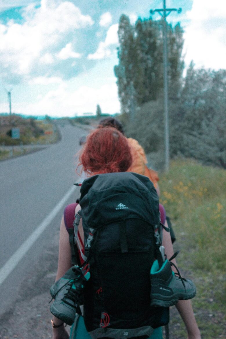Hiker viewed from behind walking on a rural road with a large backpack and shoes strapped to the outside of the pack.