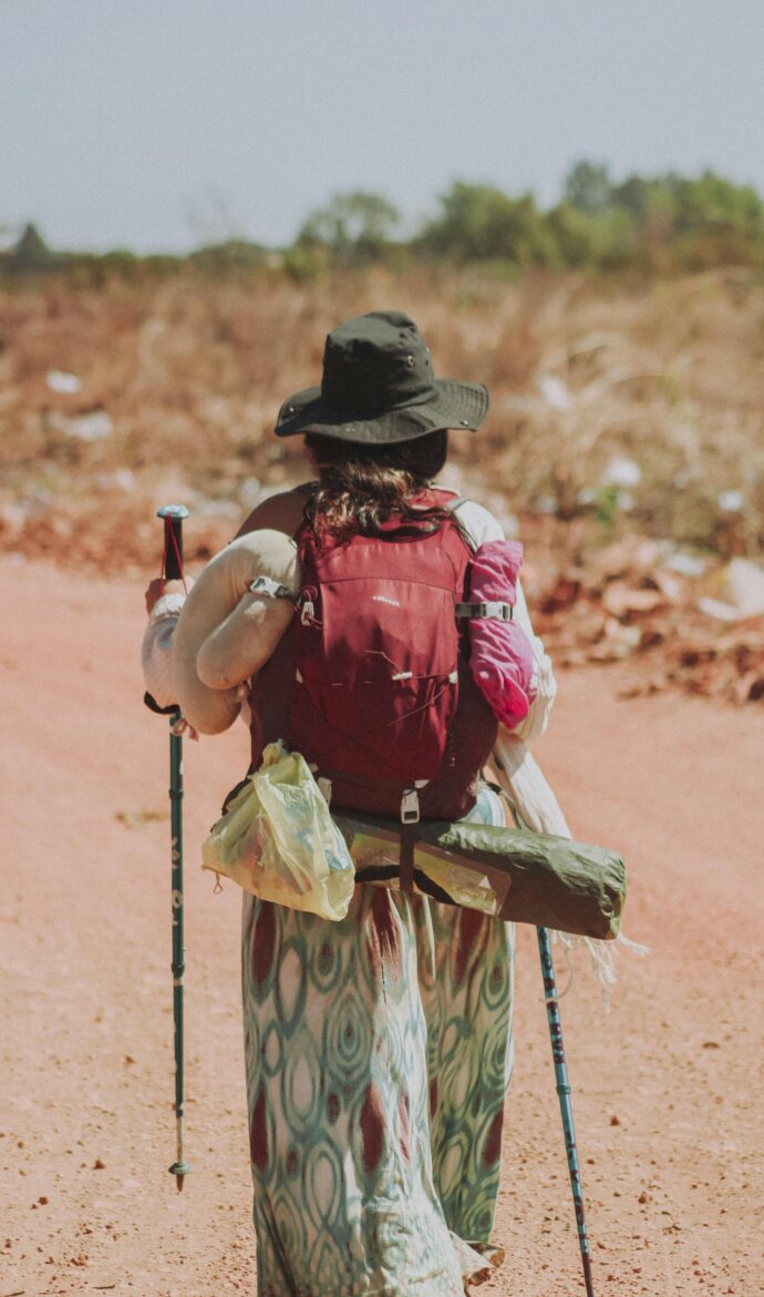 Back view of a hiker in a wide-brim hat and red backpack, standing with trekking poles in a dusty, arid landscape.