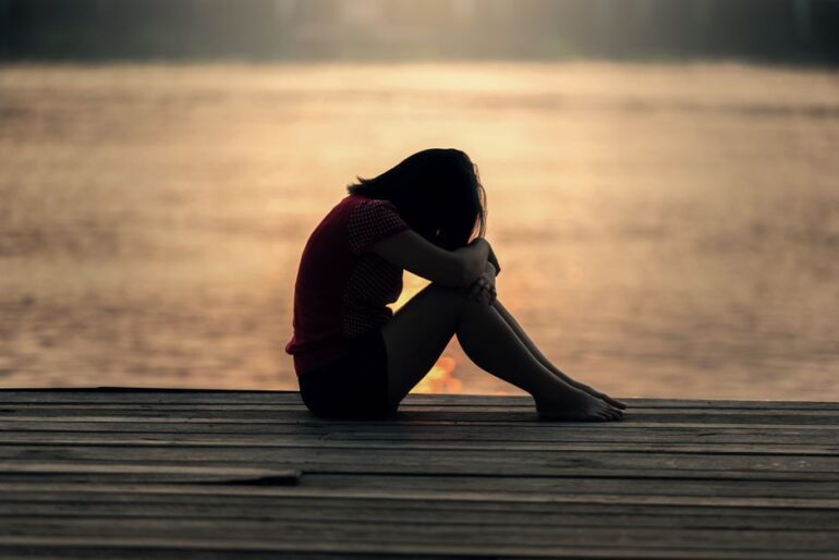 Silhouette of a person sitting on a wooden dock at sunset, head bowed and knees drawn up beside a calm lake.