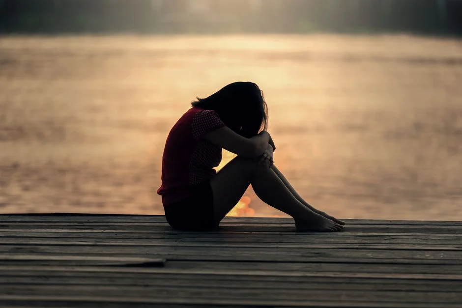 Silhouette of a person sitting on a wooden dock at sunset, head bowed and knees drawn up beside a calm lake.