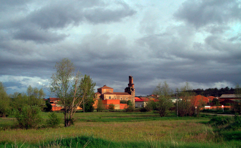 A rural village with brick buildings and a tall church tower behind green fields under a cloudy sky.