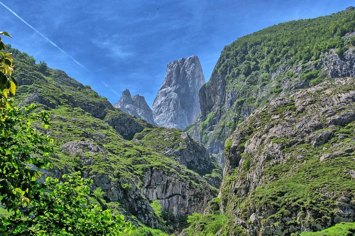 Jagged mountain peak rising between green, rocky slopes under a bright blue sky with a contrail overhead.