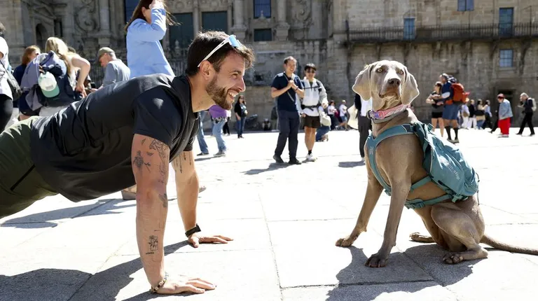 Man doing a push-up facing a wearing-dog with a teal backpack in a sunny plaza; spectators in the background.
