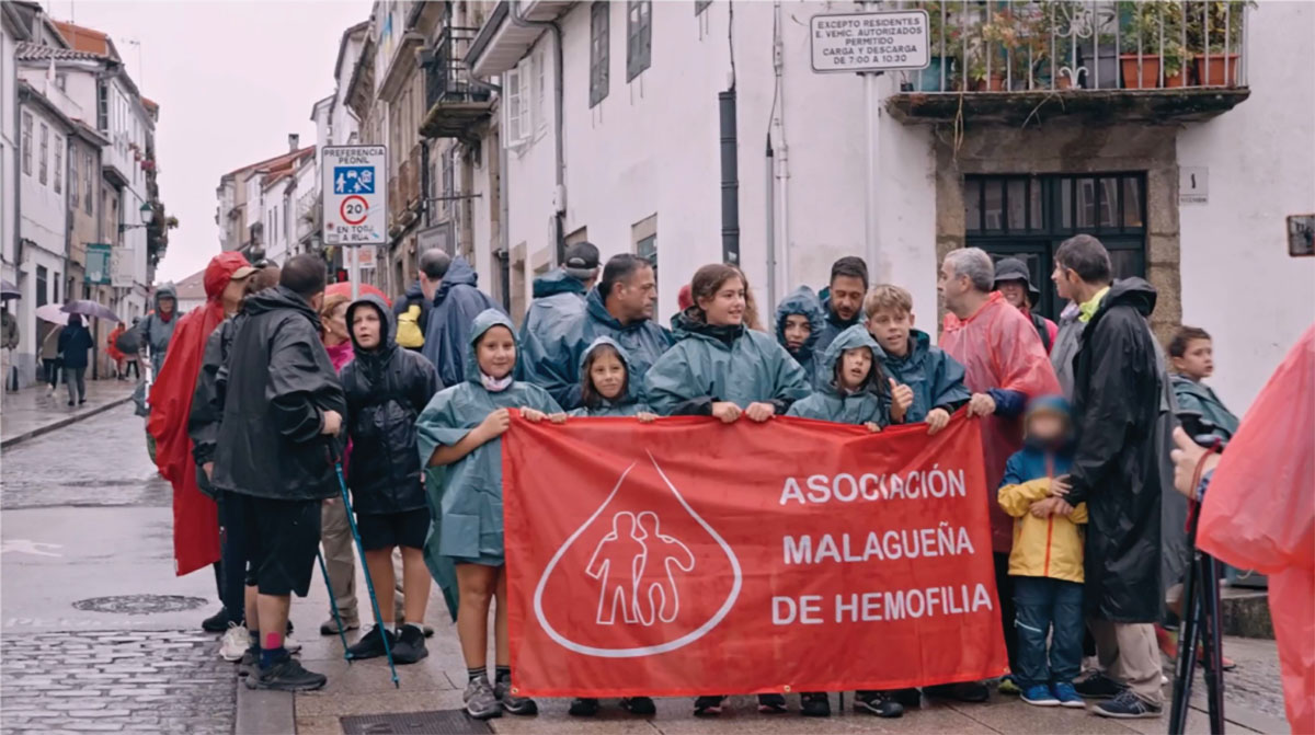 People in rain ponchos hold a red banner for la Asociación Malagueña de Hemofilia on a cobblestone street parading together in the rain.