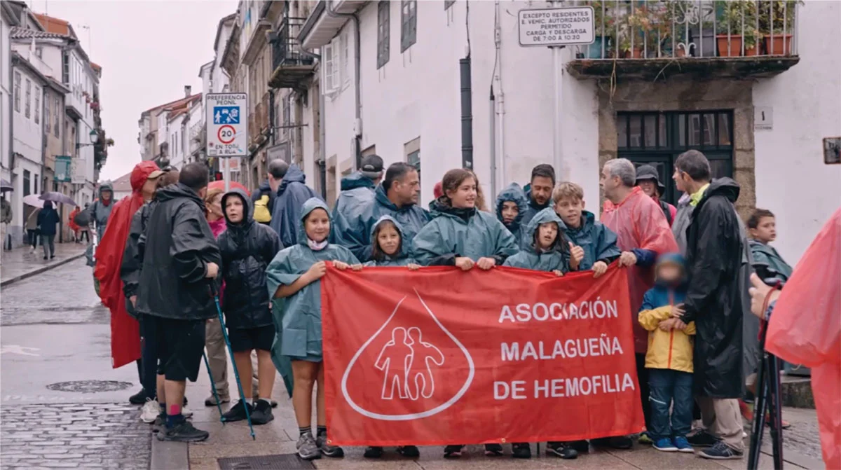 People in rain ponchos hold a red banner for the Malagueña Hemophilia Association on a cobblestone street standing together in the rain.