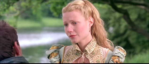 Young woman with blonde hair in an ornate period dress speaks with a man outdoors by a lake.