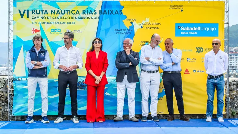 Seven people posing on a stage in front of a blue-yellow banner for the VI Ruta Náutica Rías Baixas event.