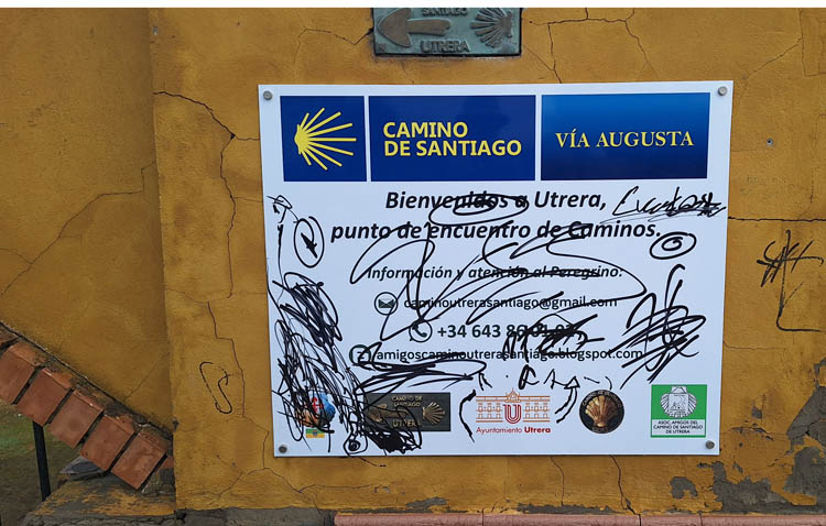 Signboard showing Camino de Santiago and Vía Augusta routes mounted on a yellow wall, with graffiti scribbles across the poster.