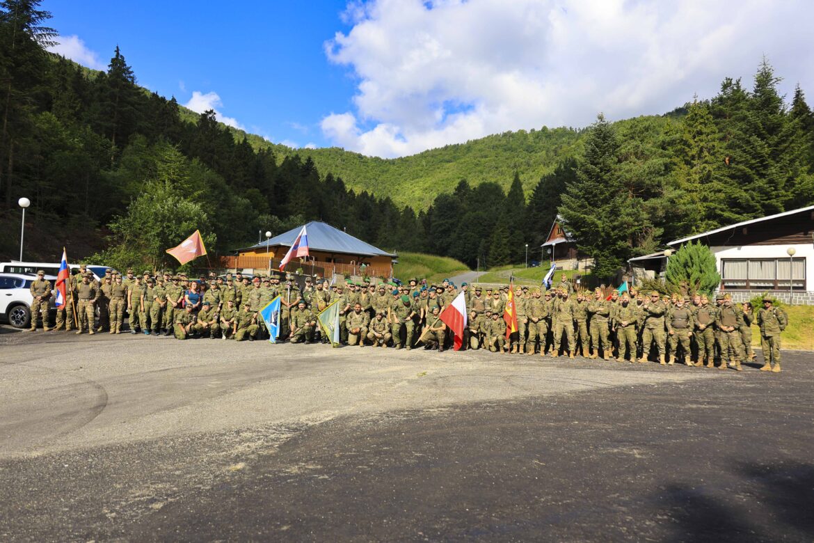 Group of soldiers in uniform posing with flags outdoors in a forested mountain setting during the day.