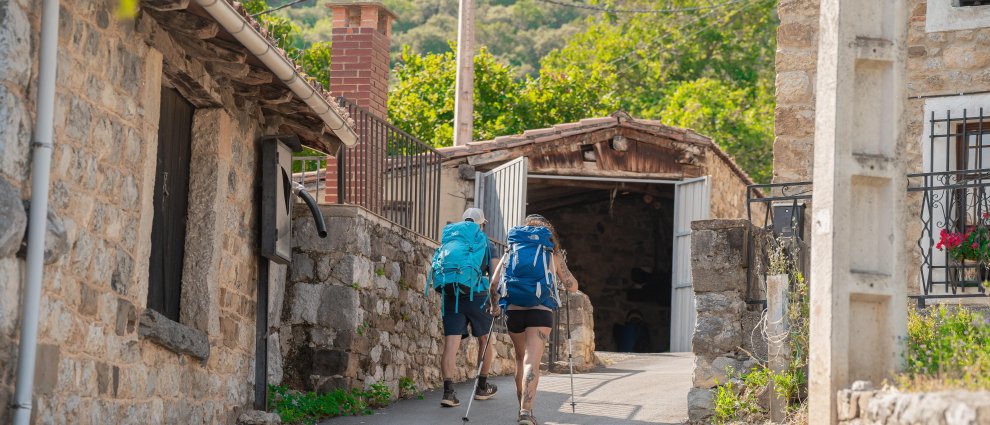 Two hikers with blue backpacks walking down a stone village street toward a shed-like building on a sunny day.