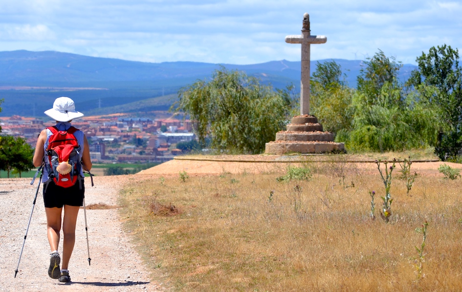 Backpacker with a red backpack walking a dirt path toward a stone cross on a hill, with a town in the valley behind.